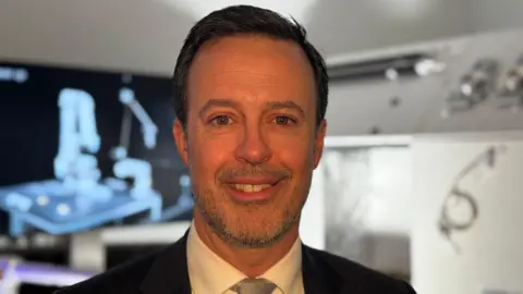 Janine Machin/BBC A head and shoulders photo of Kevin Bonavia who is smiling and looking at the camera. Behind him the background is blurred and out of focus. It appears to be some sort of science laboratory or equipment behind him.