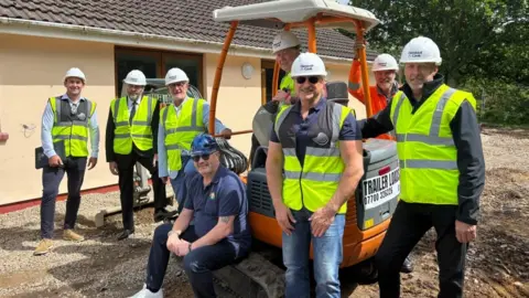 A group of men wearing hard hats and hi-viz jackets standing around a mini digger and smiling at the camera