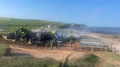 Fire fighters are seen on walking around the outskirts of the debris which is blackened and crumbling.