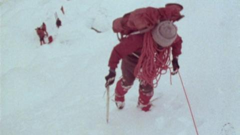 Climber in red with climbing sticks and rope in hand ascending towards the camera.  Other climbers behind further down the slope which is entirely snow.