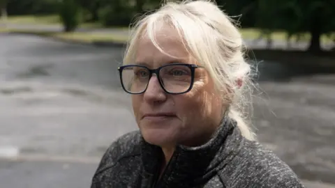 A woman with short blonde hair is looking into a camera. She has black-rimmed glasses on and is semi-smiling with a look a fondness etched on her face. She has a black zipped top on.