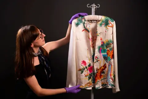 David Parry/PA Media Assignments A curator holds up a white silk top with embroidered flowers