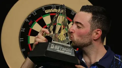 Luke Humphries kissing a pyramid shaped trophy after winning Premier League Darts