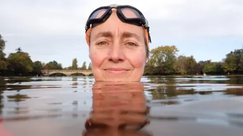 Laure Latham's head is sticking up above the water in a lake or river with a bridge and trees in the background. She is wearing a swimming cap and large black-rimmed goggles. 