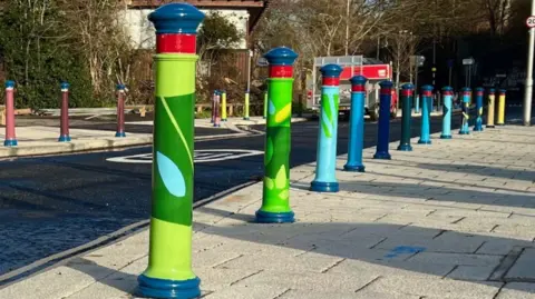 Marcus Whitehouse A row of slim bollards on a pavement, all painted in different colours, mainly green, blue, turquoise and red, some with leaf patterns 