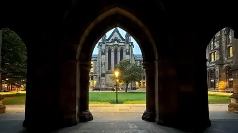 Shanti Nelson A photograph taken from inside a dark stone archway, looking out onto a courtyard with green grass and a lit lamppost. Framed by Gothic-style architecture, a large stone building stands in the background.