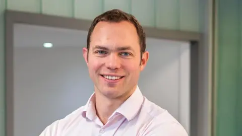 PA Media Dr Will Sedley has his arms crossed and is looking directly at the camera while smiling. He has short brown hair and is wearing a light pink shirt.