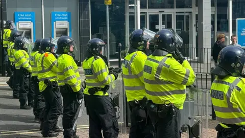 BBC Police officers with riot shields and protective helmets standing in a line separating protesters