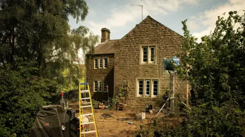 MAB Rabbit Ltd 2024 A two-storey cottage sat in the middle of greenery. Two protruding trees either side. A yellow ladder placed in front of the cottage, with industrial studio lights propped up facing the building. 