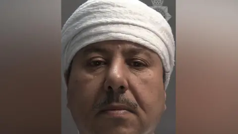 A custody picture of a man with a moustache who is wearing a white head covering. 