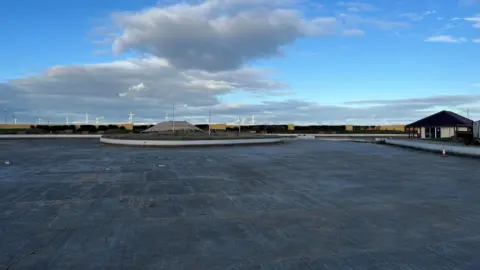 A large patch of concrete at the empty lake. Redcar's wind turbines are visible in the distance. the Boathouse cafe is on the right side of the lake.