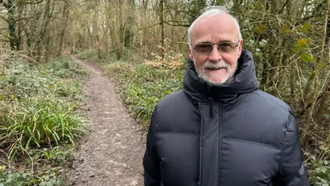 Peter Martin is wearing a dark puffer jacket and has short greying hair. He is wearing tinted sunglasses and is smiling at the camera. He is standing in a woodland.