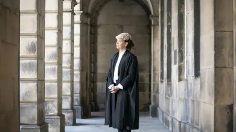 PA Media Lord Advocate Dorothy Bain - a woman with dark curly hair tied back behind her head - pictured in the middle distance, standing with her hands clasped, between the stone pillars of Edinburgh's Court of Session, dressed in the traditional outfit of an advocate - long black robes over a white shirt, with a lawyer's wig