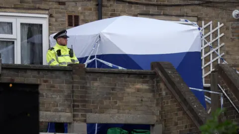 PA Media A blue and white police tent and a police officer standing outside a house