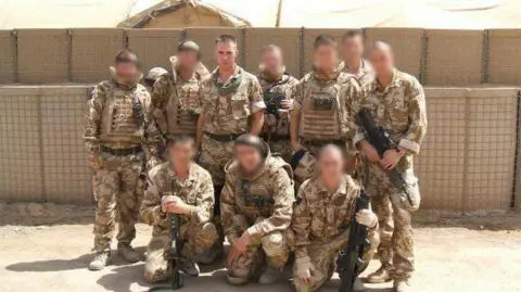 Chris Draper Two rows of soldiers, some armed with rifles, posing for a photo wearing desert camouflage uniform with a sand-coloured protective wall behind them. Nine of the ten faces have been blurred and only Chris Draper is identified