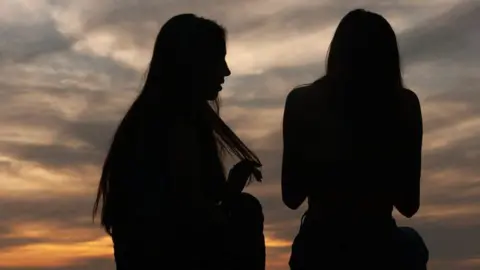 A silhouette image of two young women. They have long hair.