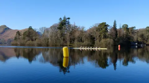 Ian Gordon Side shot of a single yellow rowing boat on the water. There are multiple people rowing it. Behind them are trees and hills which are reflected in the crystal clear water. There is a yellow and orange buoy in the photo. 