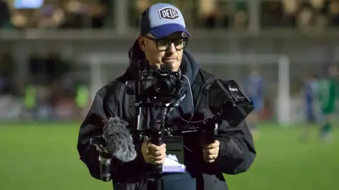 Rich Phippen - a man with a blue hat on holding a camera with a microphone on, while standing on a football pitch. 