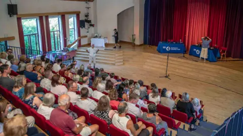 An auditorium with an audience seated in rows and steps leading to a stage which is hosting Gardeners' Question Time.