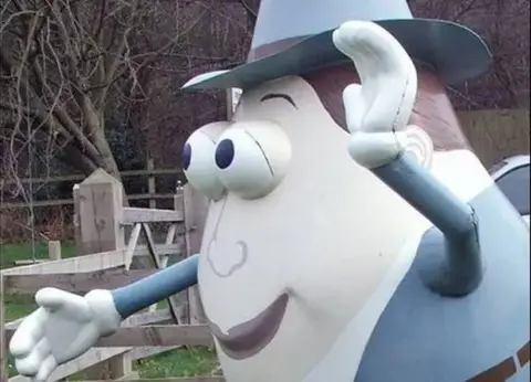 A large statue of the children's toy, Mr Potato Head, stood beside a wooden fence. The statue is not in the traditional Mr Potato Head colours, he is painted in off-white, with a painted grey suit, hat, brown hair and brown lips.