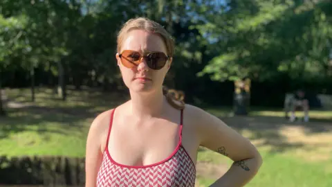 Owen Sennitt/BBC A woman wearing a red tank top with sunglasses while standing outside. Her hair is in a middle parting and braided at the back. She has one hand on her hip and is looking at the camera. Behind her is some grass and trees 