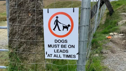 A sign attached to a fence  tells people that "Dogs must be on leads at all times".
There is a wooden style in the background and a stone gatepost in the foreground. 