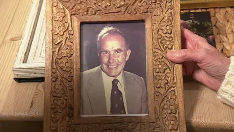 A person holds a framed photo of a man. The man is smiling at the camera. He has short grey hair and wears a grey shirt with a tie. 