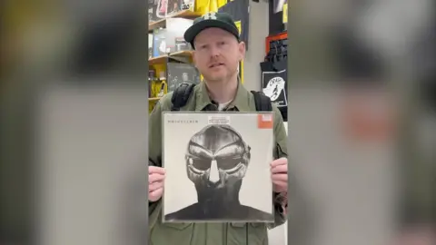 A man in a green shirt and cap holds a record called Madvillainy