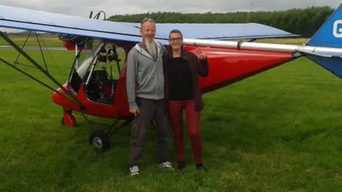 Handout A man and a woman stood in front of red glider plane sited on a patch of grass. The man on the left is wearing brown trousers and a grey zip-up hooded top. He has a long grey beard. The woman, on the right, has dark hair and is wearing glasses. She is doing a V sign with her fingers and is wearing burgundy trousers, a black top and a dark burgundy jacket. 