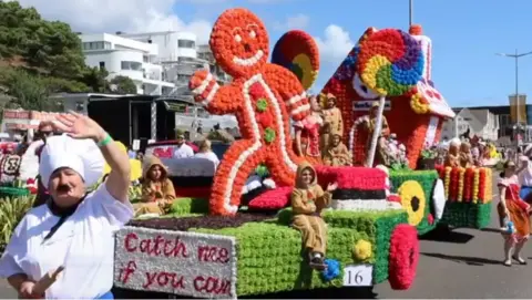 A Battle of Flowers float parades down the avenue. It depicts a gingerbread man running, with a lollipop house seen in the background