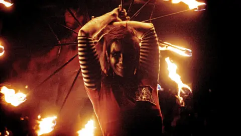 A woman is looking down the camera and is spinning fire on sticks above her head. She is dressed in black and white clothing and has her face painted white with black stars around her eyes