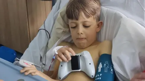 NHS Blood and Transplant A child sits upright in a hospital bed surrounded by medical equipment, with a blood transfusion bag and monitoring devices visible above and beside the bed. 