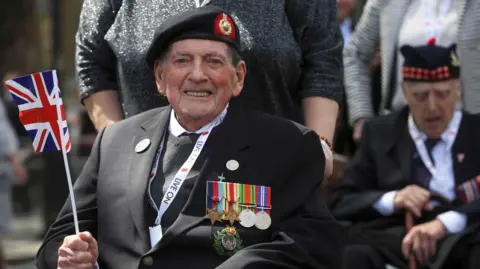 A veteran waves a Union Jack flag as he takes part in a VE Day Parade. He is wearing a suit with a line of medals across his chest.
