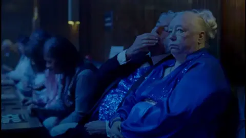 Taylor Swift - Opalite The image shows an elderly couple sat together in a dimly lit space, they both have bored expressions and are dressed in royal blue ballroom dancing outfits. 