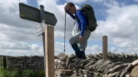 A man climbing a gate at a brick wall in the lake district. He is holding a walking stick and has a back pack. The backdrop is a blue cloudy sky.