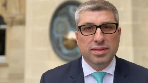 West Northants Council leader Mark Arnull wearing a navy suit, white shirt and turquoise tie