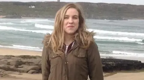 Natalie McNally, with long blonde hair, wearing a dark green coat. She is standing on a beach with the sea visible behind her. 