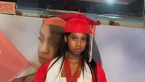 Melissa Dedeaux Nikyra Cheyenne Dedeaux mengenakan topi dan gaun wisuda berwarna merah. Gambar close up, terlihat tepat di bawah bahu.