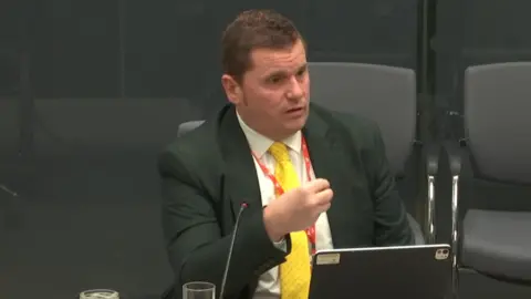 Andrew Morgan sat at a desk speaking at a committee. He is wearing a dark suit with a white suit and a yellow tie. He has a computer and microphone in front of him, and is gesturing with his right hand.