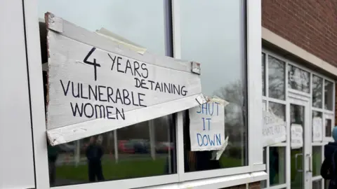 Close-up of windows of a detention centre. The windows are plastered with handwritten signs stating '4 years detaining vulnerable women' and "Shut it down'.