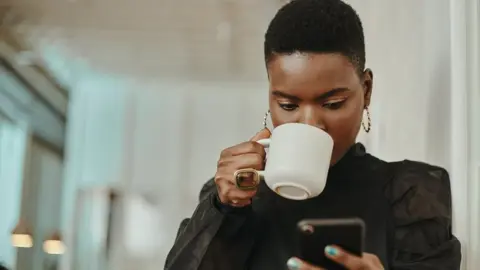 Getty Images Woman drinking a hot drink and looking at her phone