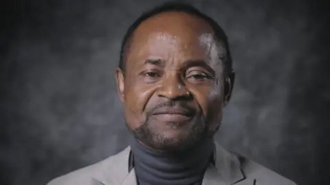 Head shot of Agomoh Paul from BBC interview, wearing grey polo neck and jacket