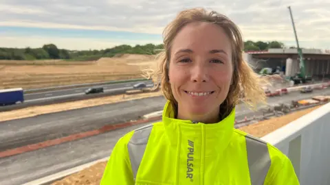 A woman with wavy, shoulder-length blonde hair smiles as she stands in front of a huge construction project on an A road. There are cranes and large vehicles parked along the road and lots of sand on either side. She is wearing a yellow hi-vis jacket with silver strips on it.