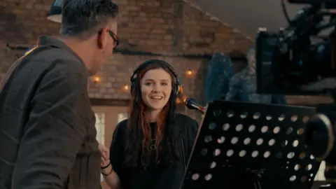 Tourettes Action Grace Cope stands behind a microphone and camera. She is wearing wearing headphones and smiling at a man in the foreground who is side-on to the camera. A music stand can also be seen, and there is a brick wall behind her.