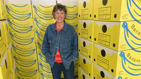 Lady smiling with short wavy hair, blue denim jacket, striped red and orange jumper and blue jeans. She is stood next to a stack of yellow boxes with Bananarchy written on them.