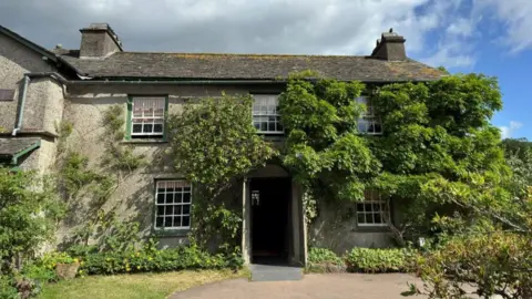 National Trust Hill Top House near Hawkshead. It is a two-storey cottage with sash windows. There are creepers up the walls, with a high concentration of green leaves to the right side of the property. There are shrubs and flowers surrounded the house.