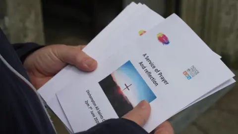 An order of service was for the event with the words "A Service of Prayer And Reflection" written on the front. There are a number of them being held by a pair of hands. 