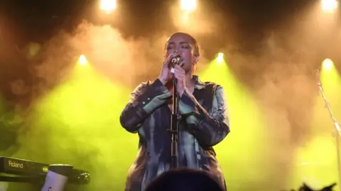 A woman on stage, singing into a microphone, with yellow lights and smoke in the background. The singer is holding the microphone with both hands and wearing a black and grey dress. 