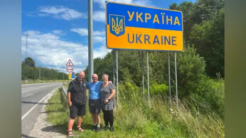Three people - two men and a woman - stand in front of a large sign that says Ukraine in Ukrainan and English on a large blue and yellow background. The sky behind them is blue and the verge is a lush green colour.
