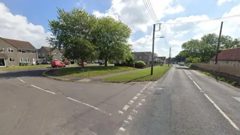 Google A screenshot from Google street view showing a junction turning into a residential street from the main road. There is a grassy patch in the middle with two mature trees, and a telegraph pole with wires stretching across the road.